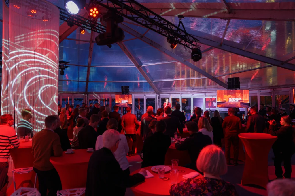 Yleisöä teltassa joka on valaistu punaisella valolla, sekä lava jossa juontaja puhuu mikrofoniin.