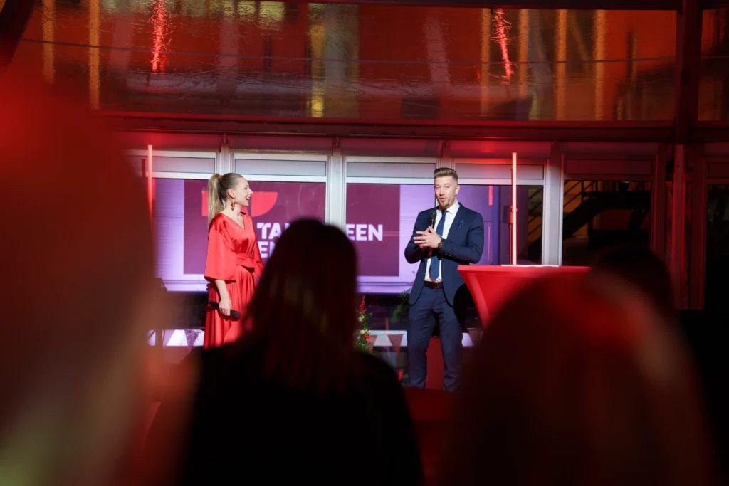 Yleisöä selin kameraan sekä taustalla lavalla juontaja Sanna Laitinen ja haastateltava miesoletettu