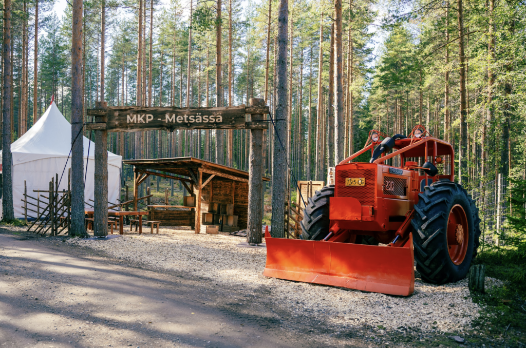 Metsässä oleva messuosasto, jossa iso punainen traktori, puinen ohjelmalava ja valkoinen teltta.